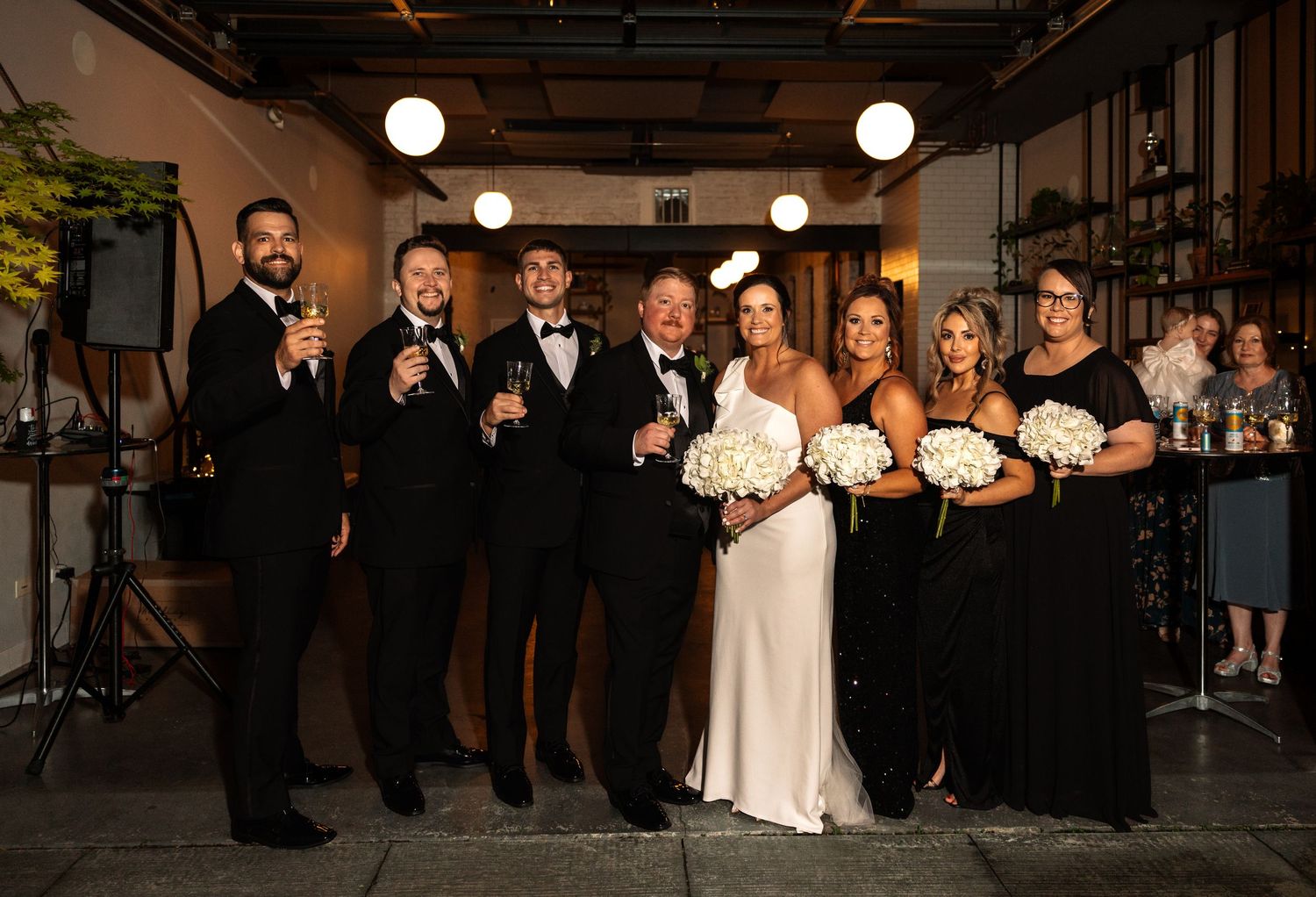 Wedding party in black formal wear poses with bouquets and champagne at indoor reception venue.
