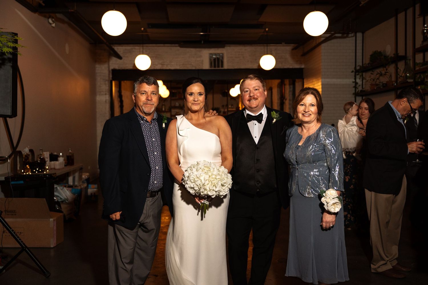 Bride and groom smile with two family members under warm lights at elegant indoor wedding reception venue.