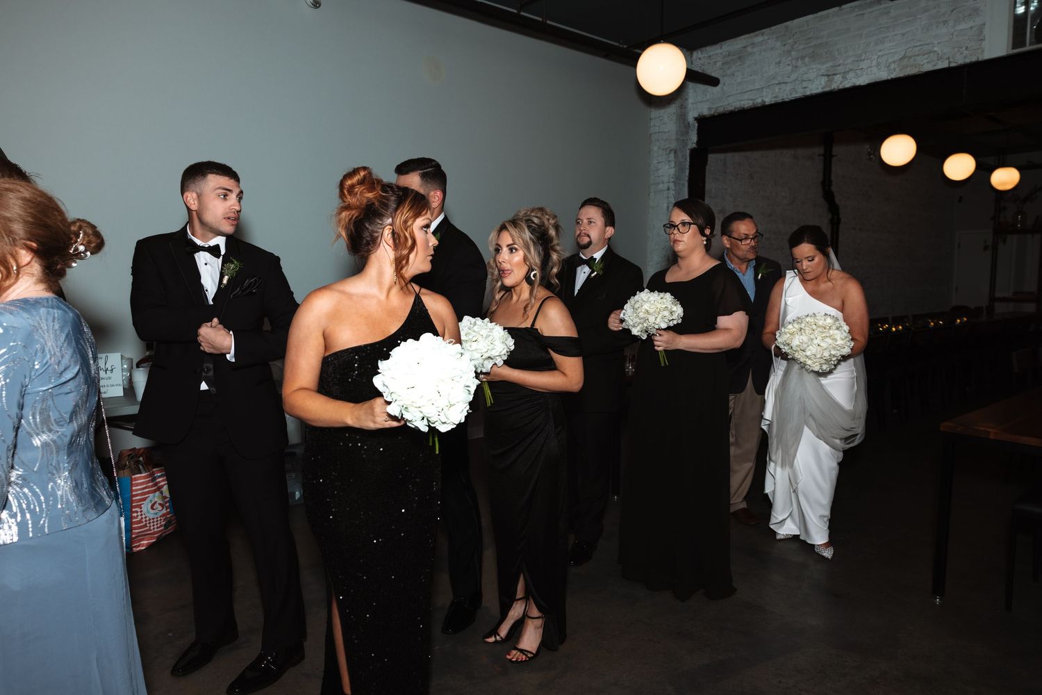 Wedding party in black formal attire holding white bouquets during ceremony procession.