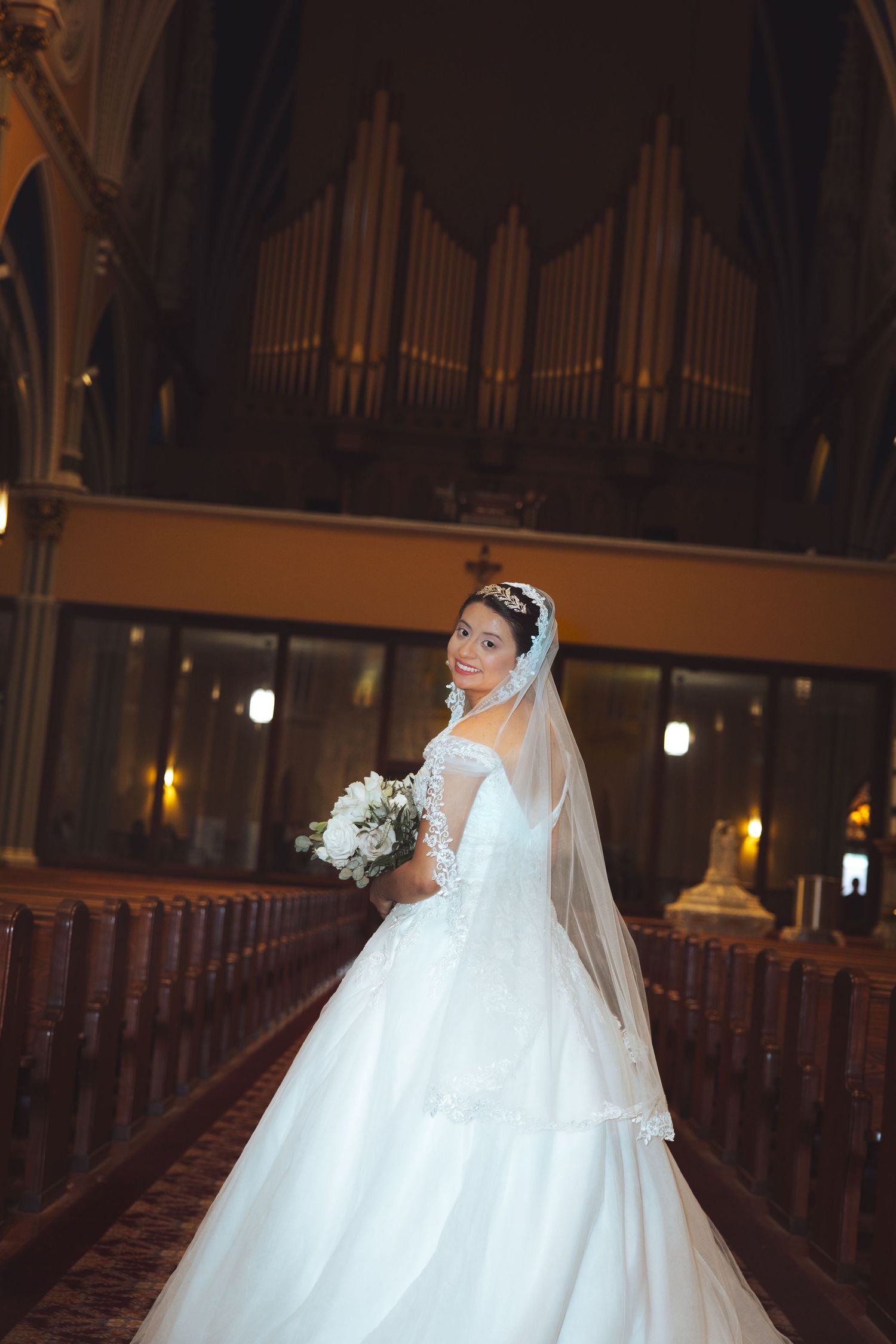Elegant bride in lace ball gown with veil and tiara poses in church aisle with pipe organ backdrop.