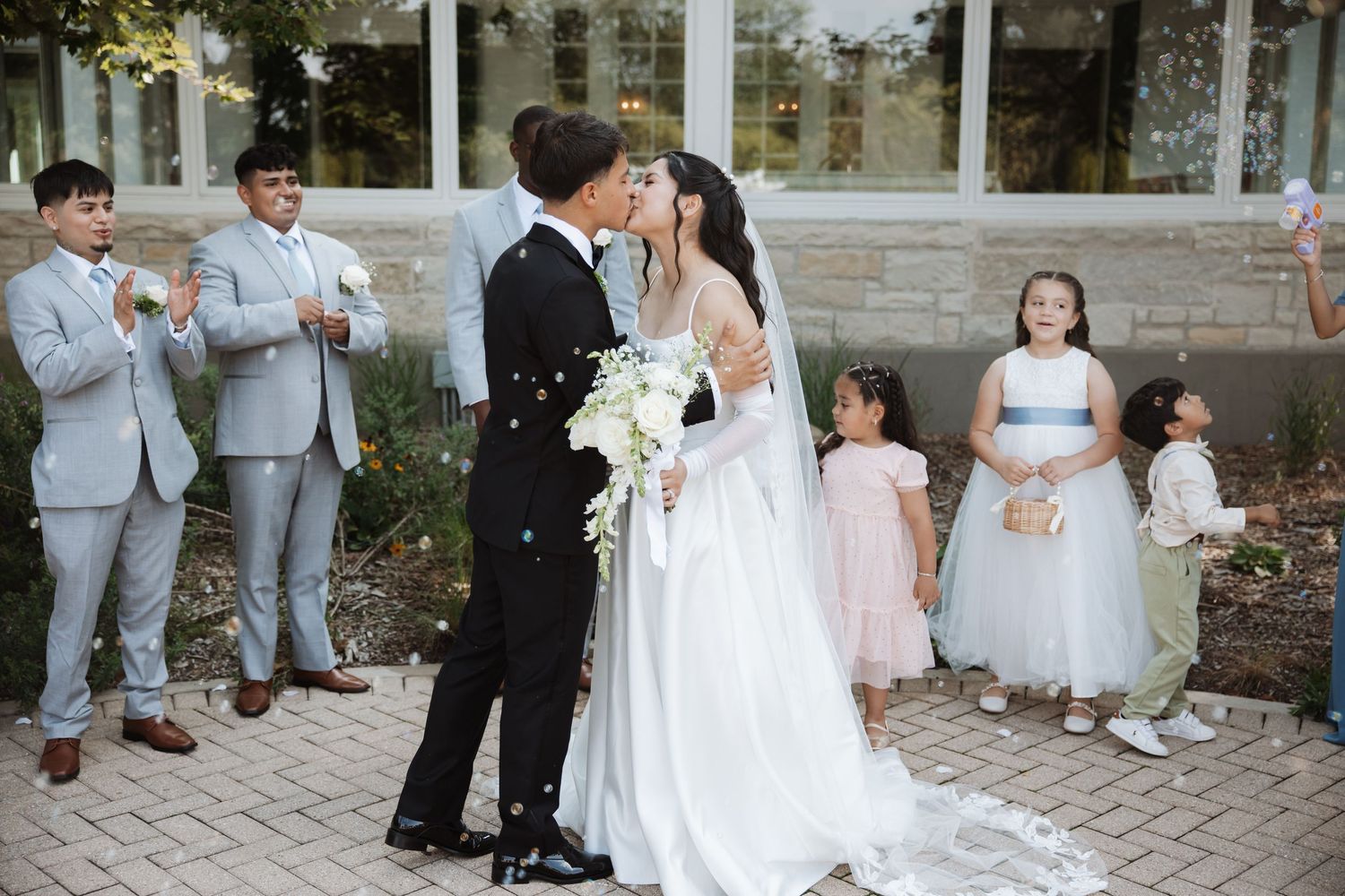 Newlyweds kiss as confetti falls while wedding party of groomsmen and flower girls celebrate outdoors.