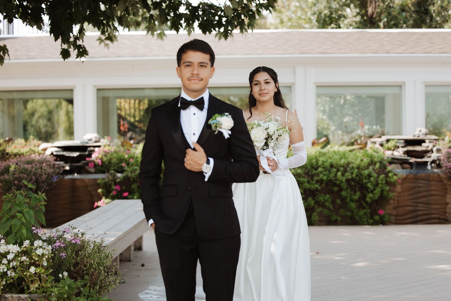 Groom in black tuxedo poses with bride in white wedding dress holding bouquet at outdoor venue.