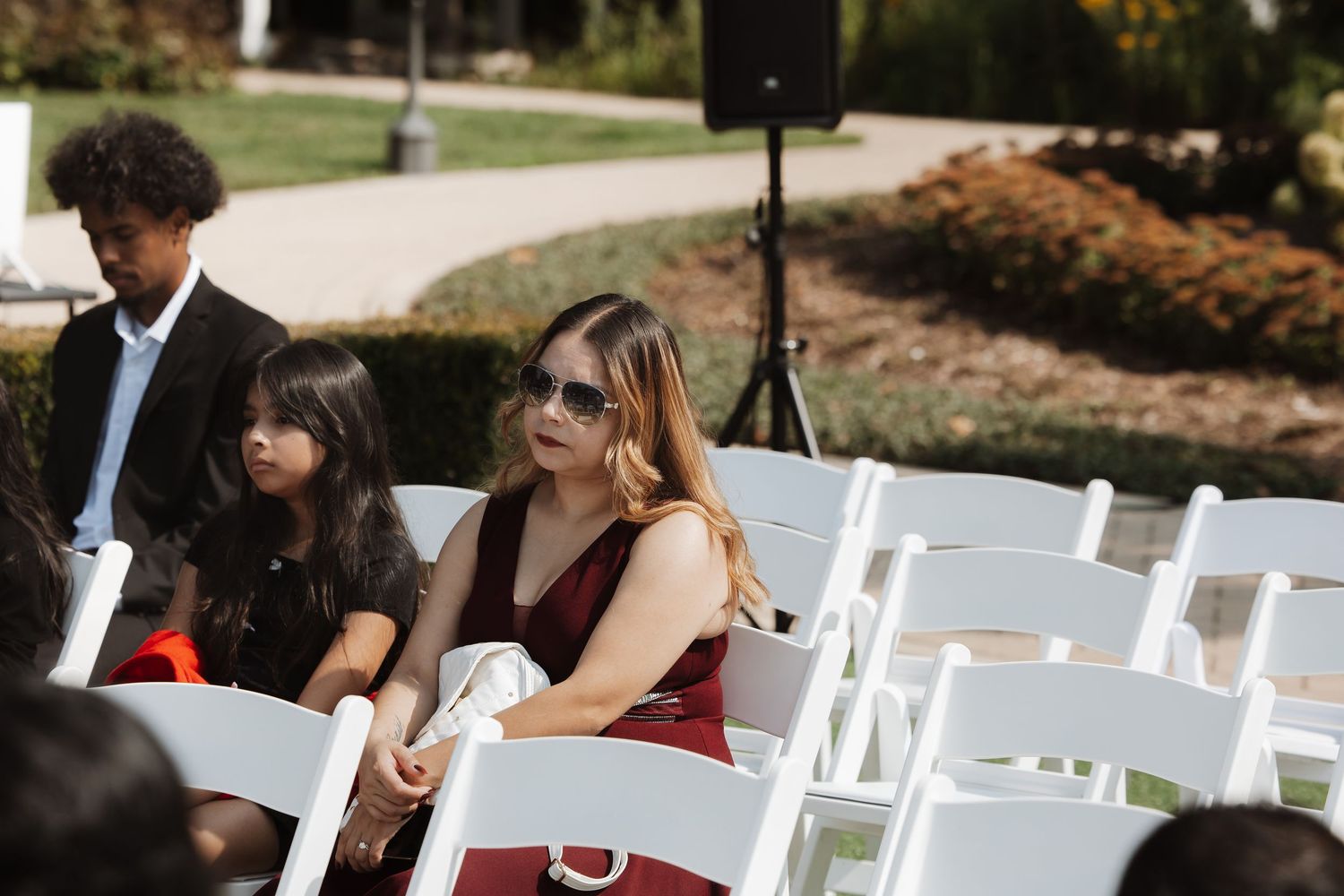 Wedding guest in red dress and sunglasses sits with children in white chairs at outdoor ceremony.