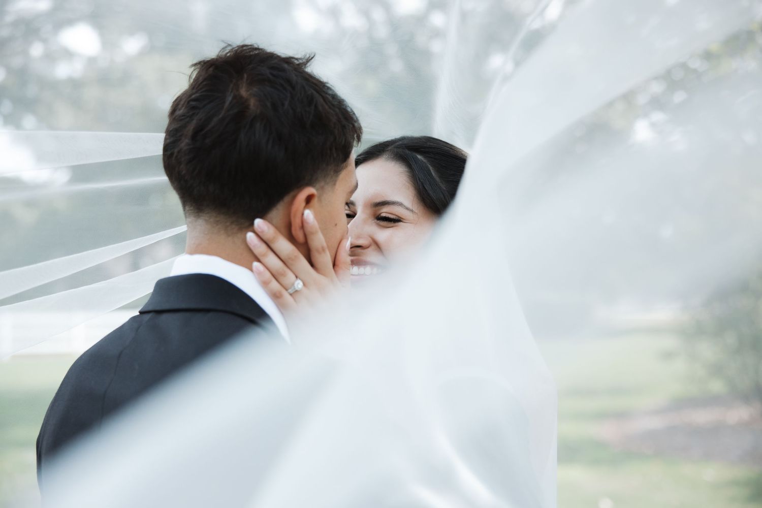 Bride laughs while holding grooms face under a white wedding veil, capturing a candid joyful moment.