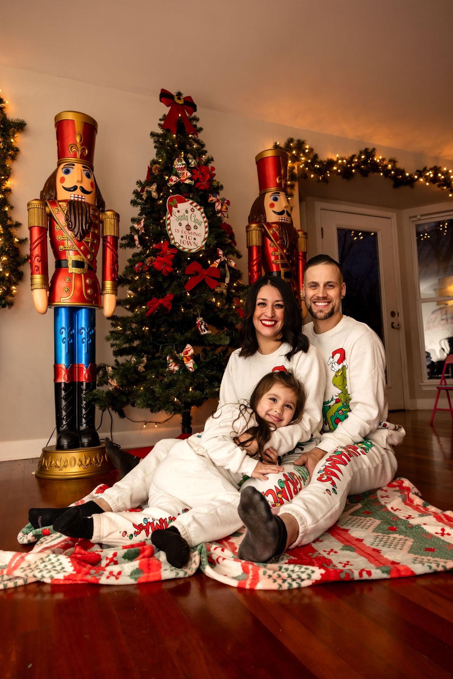 Happy family of three in matching Christmas pajamas sitting on a festive blanket near a decorated holiday tree.