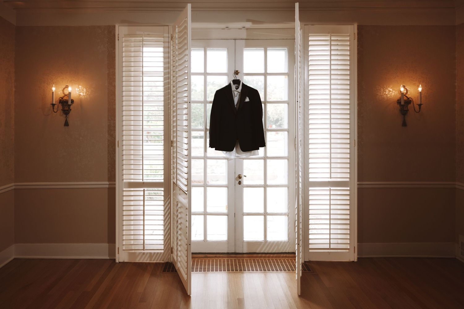 Groom's black suit hanging on French doors in warmly lit elegant room.