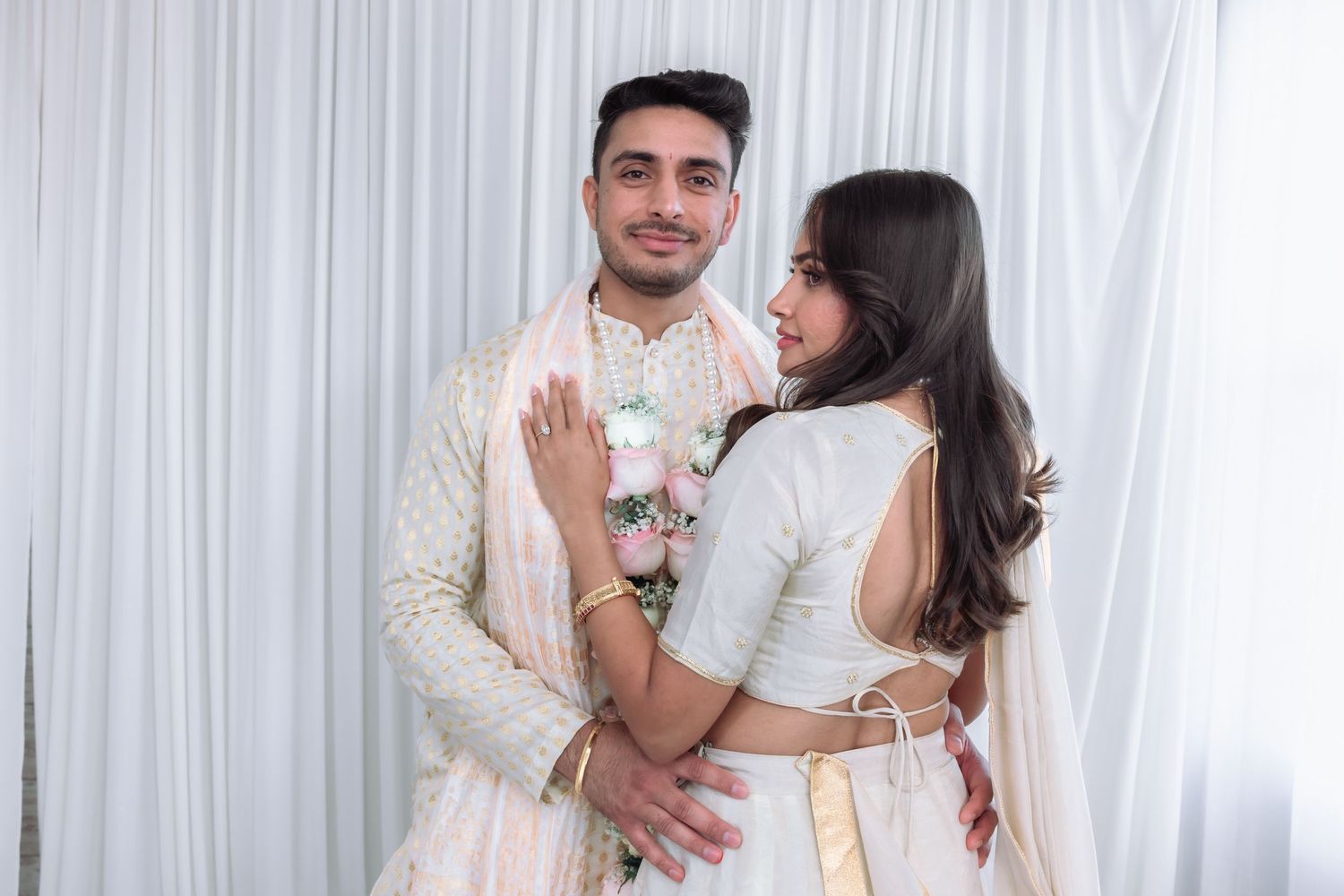 Indian groom in cream sherwani embracing bride in white lehenga during their traditional wedding ceremony.