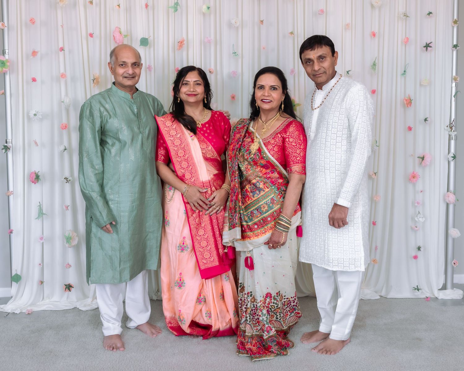 Indian family of four smiling in traditional attire, standing barefoot against a white floral backdrop.