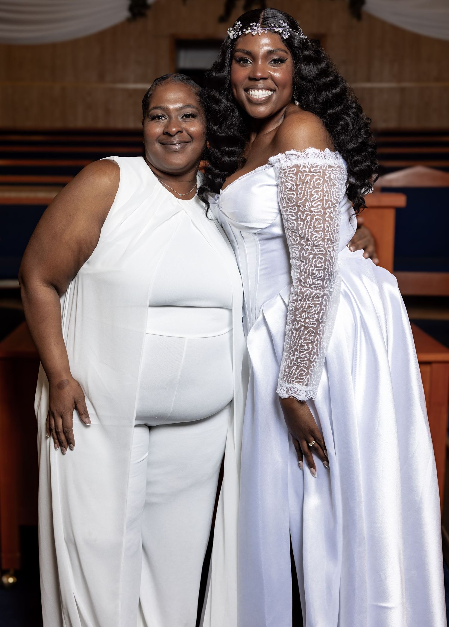 Two women in white formal attire smiling together inside a church, one in a bridal gown.