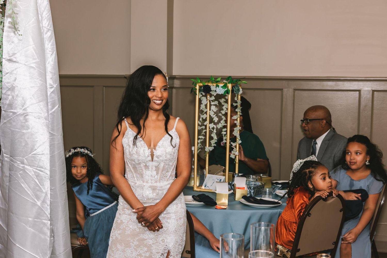 A radiant bride in a white lace gown smiles warmly surrounded by flower girls at a wedding reception.
