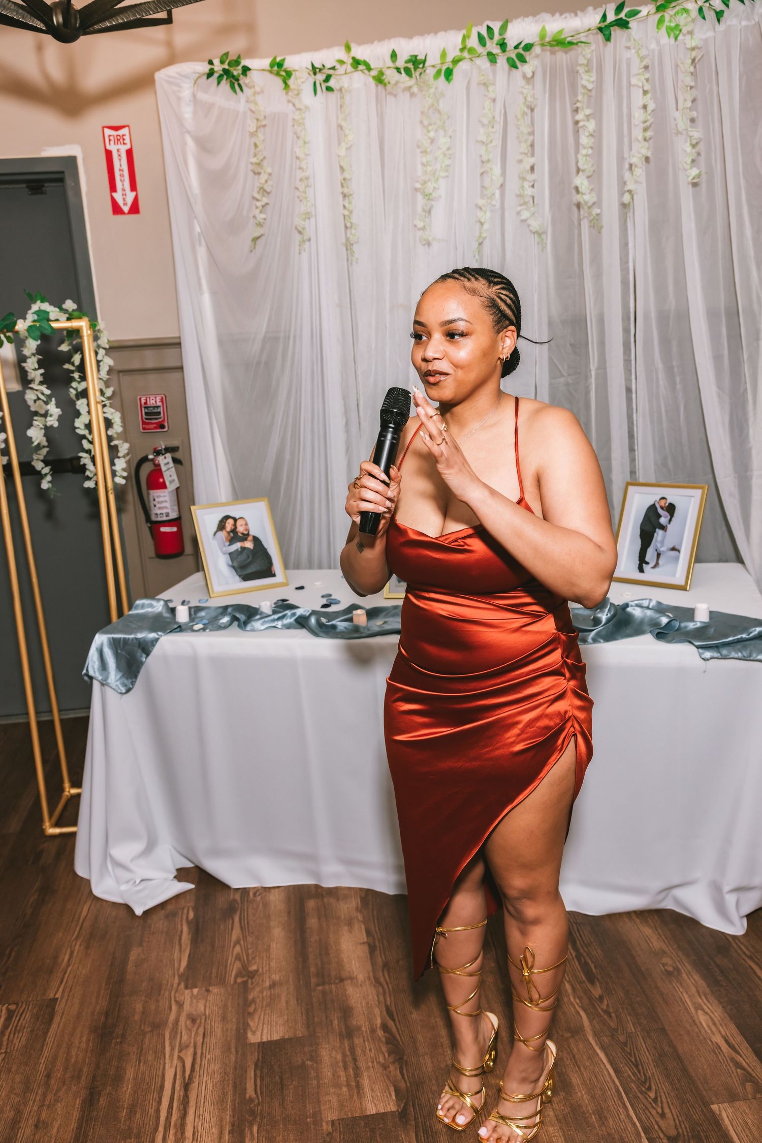 Woman in rust satin dress holds microphone while giving a speech at a wedding reception.