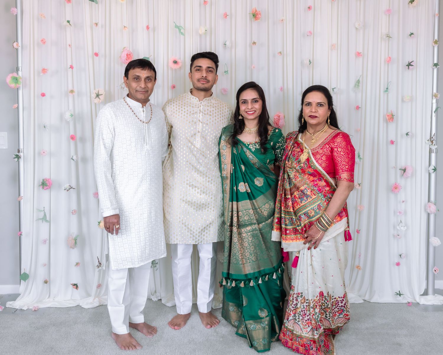 Four guests in traditional Indian wedding attire smile together against a floral decorated backdrop.