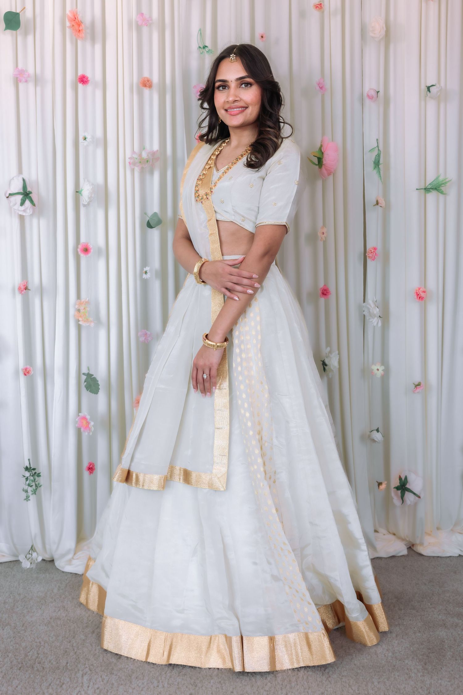 Smiling woman in white gold-trimmed lehenga posing confidently before a decorative floral backdrop.