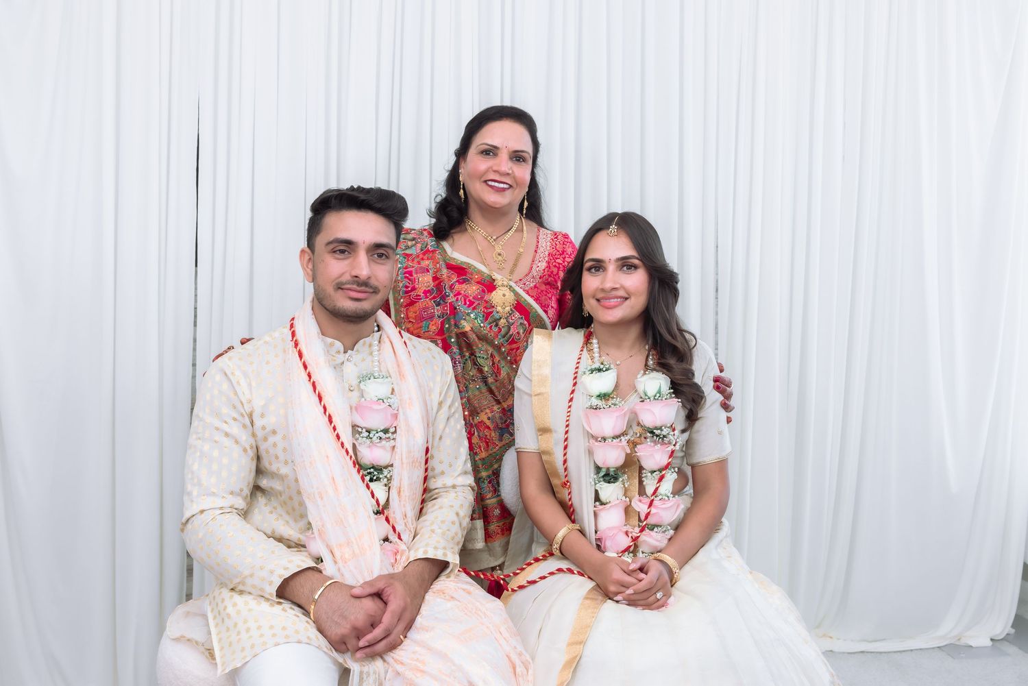 Indian bride and groom in white wedding attire posing with elder woman in pink saree for family photo.