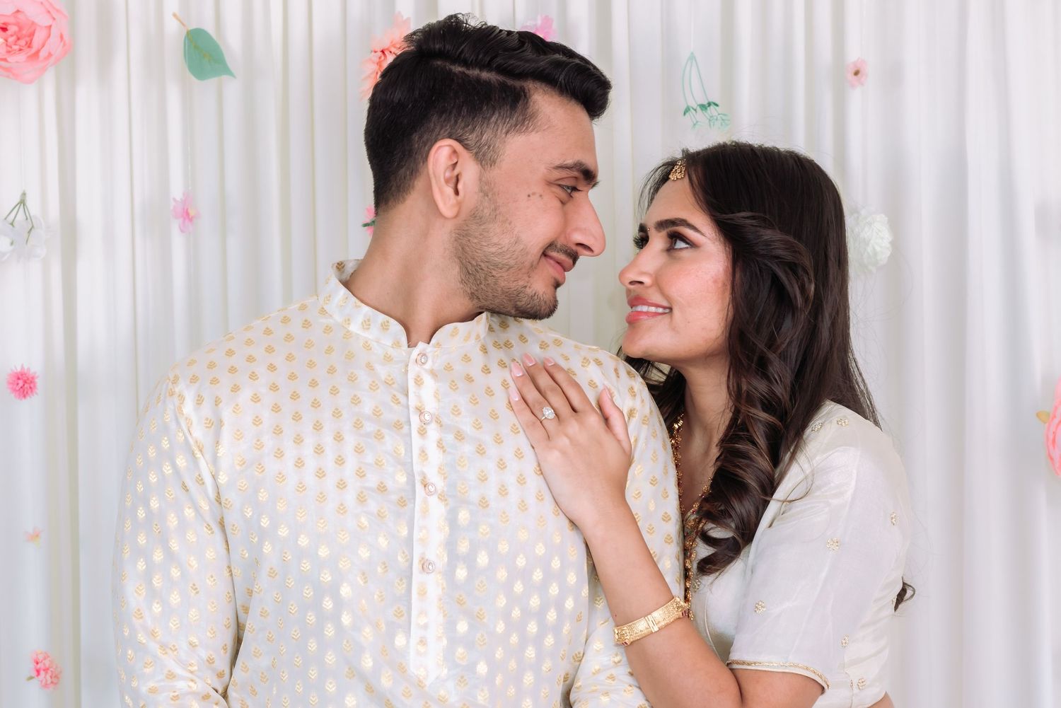Indian couple in white traditional attire sharing a loving gaze during engagement ceremony with floral backdrop.