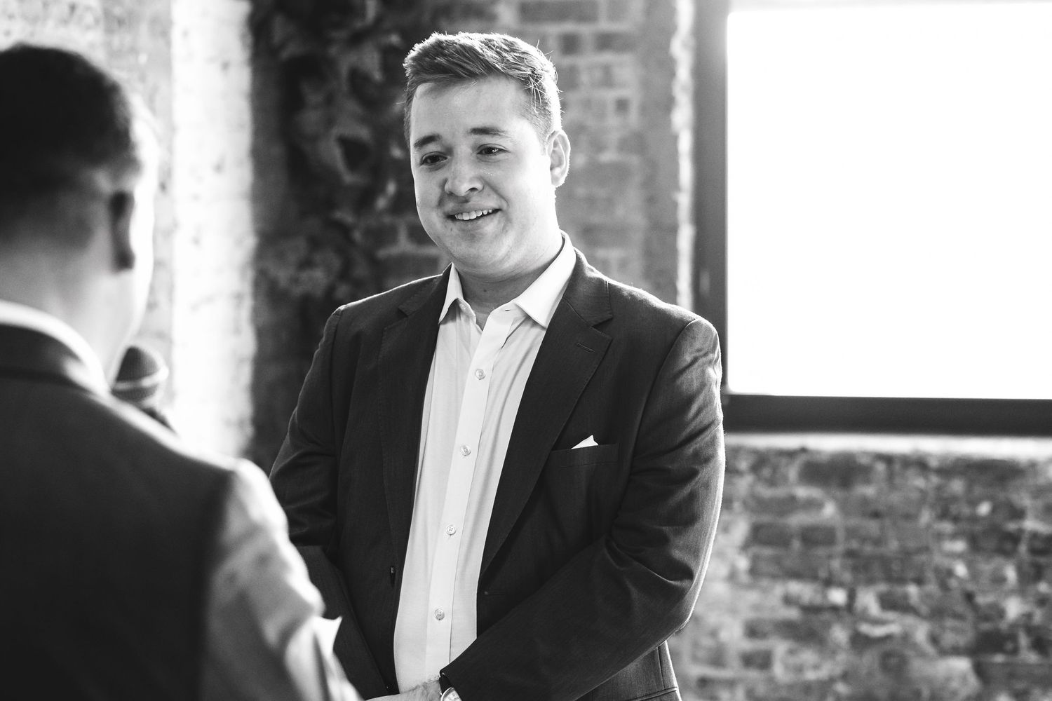 Young man in dark blazer smiling warmly during a handshake at an indoor brick-walled venue, black and white photo.