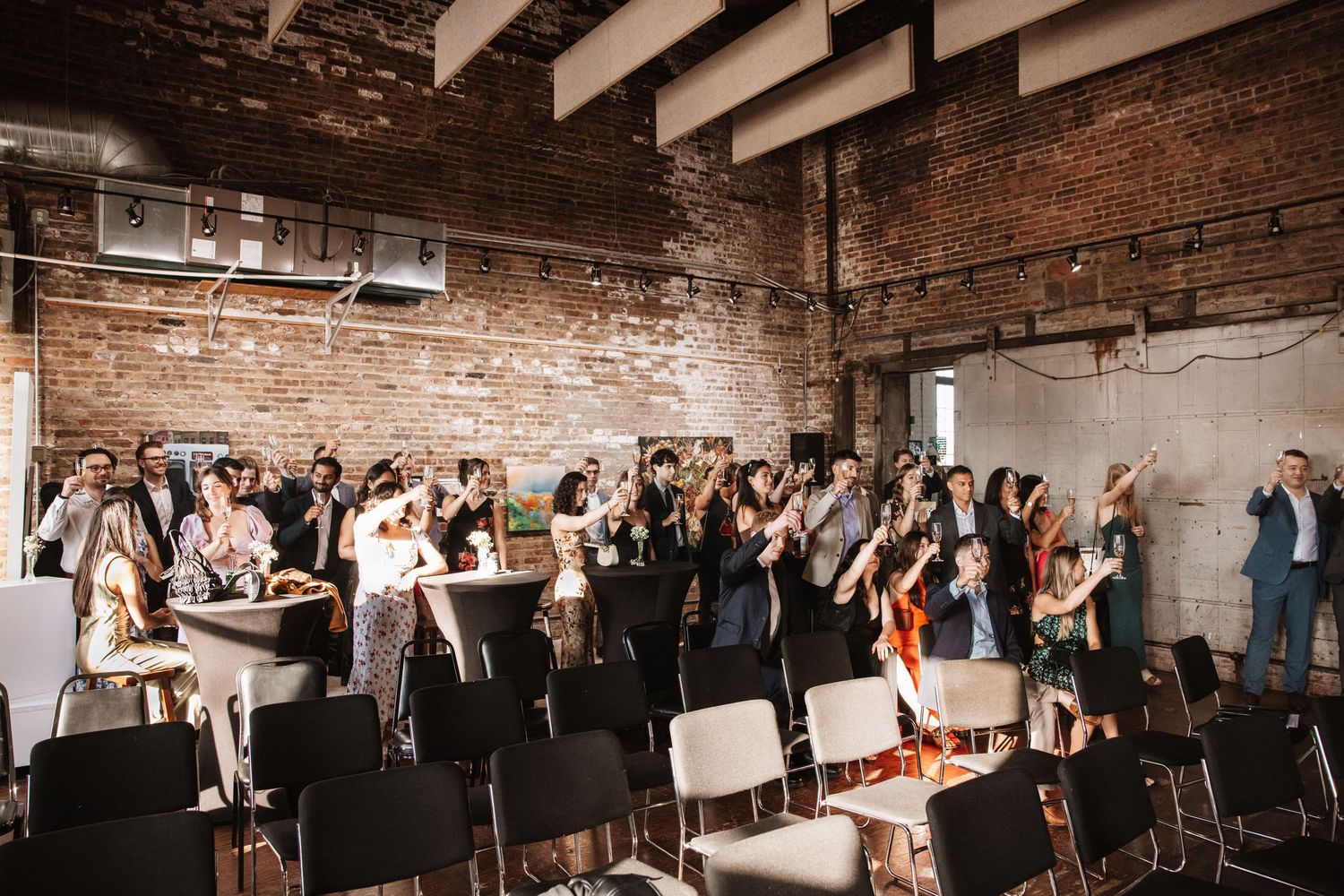 Wedding guests mingling and applauding in rustic industrial brick event space with folding chairs.