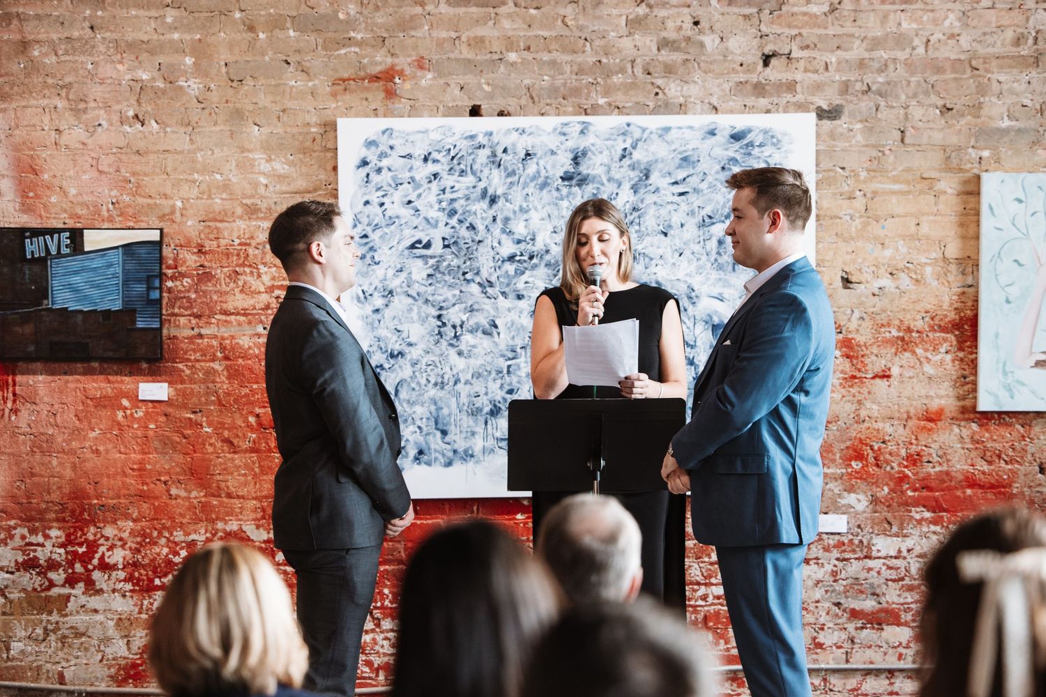 Woman officiates a ceremony between two men in suits in front of abstract wall art.