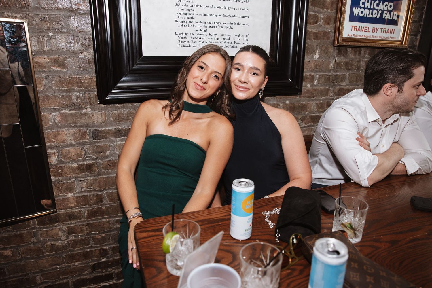 Two women in green and black outfits posing at a bar table.