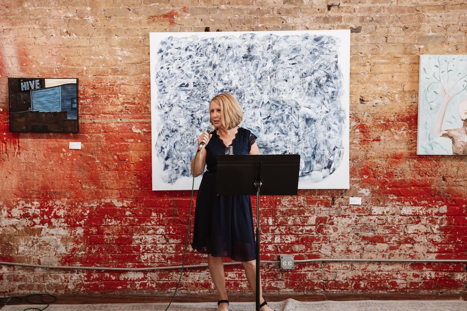 Blonde woman in navy dress presents at podium before large blue abstract art on brick wall.