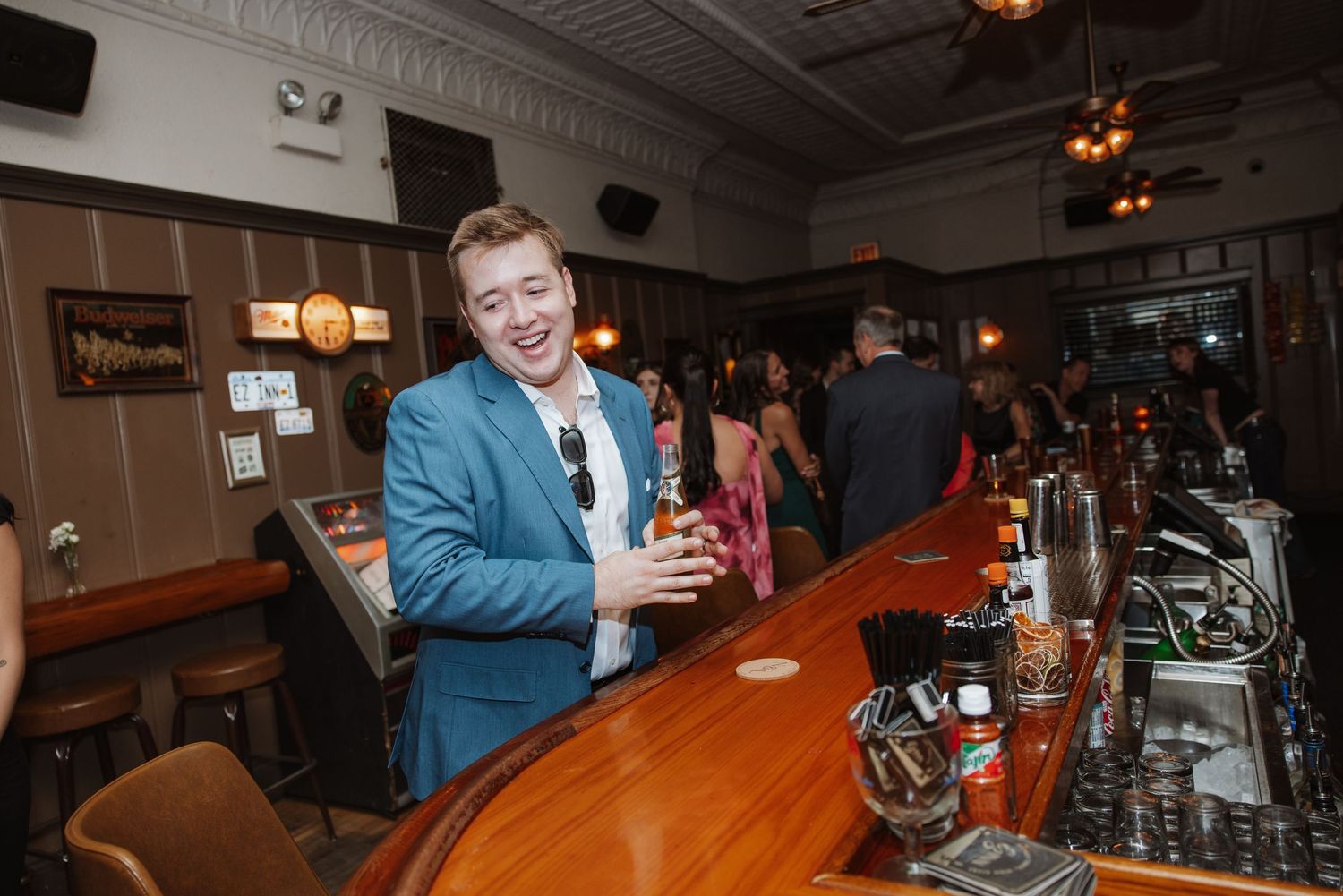 Man in blue suit smiling at bar holding beer bottle at evening event.