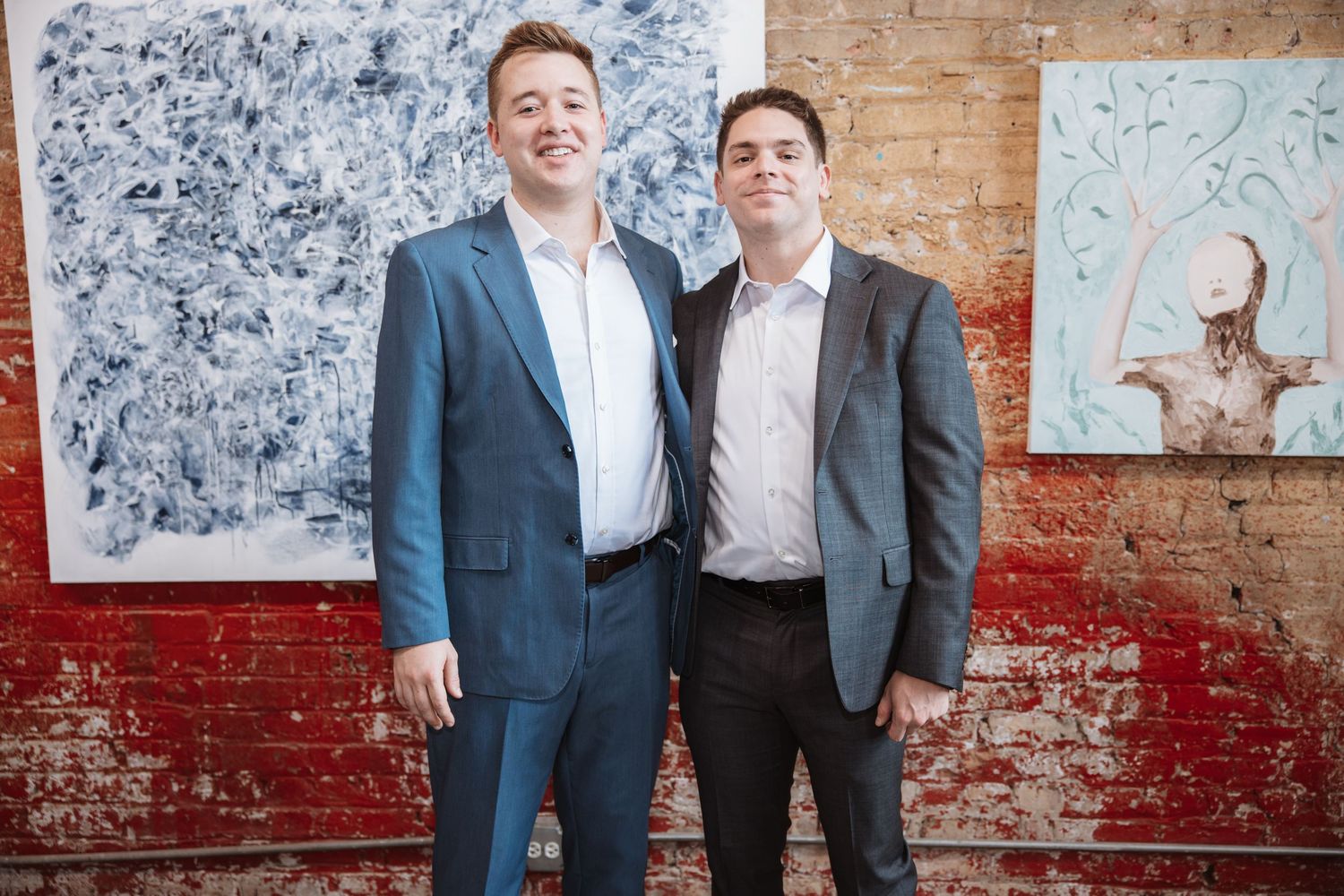 Two men in suits smile together at an art gallery event with brick wall backdrop.