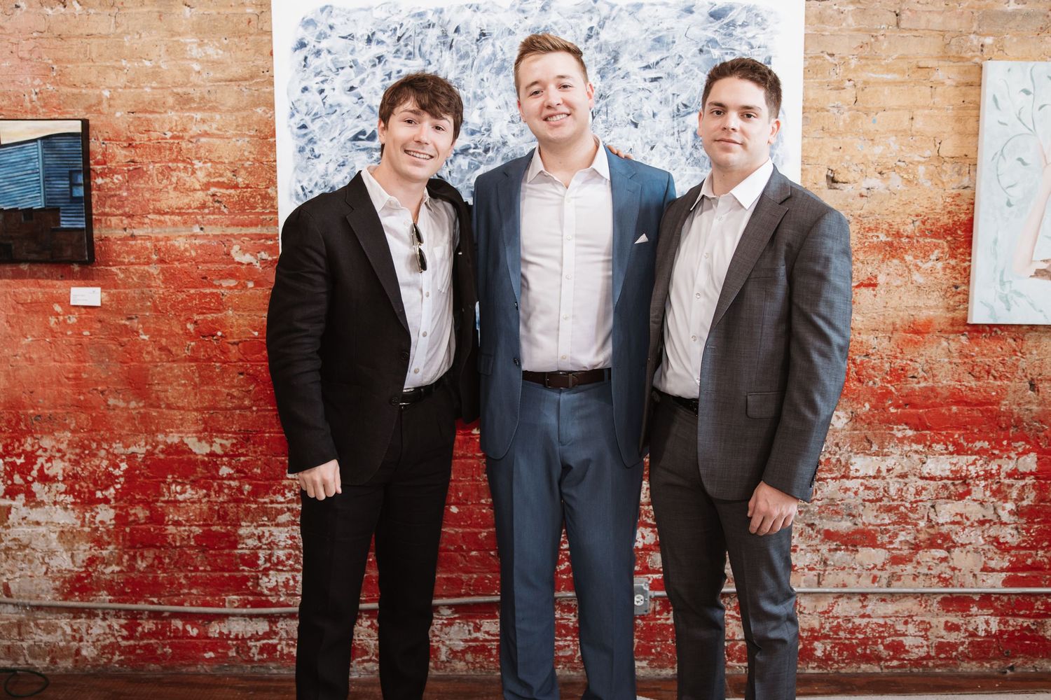 Three men in formal attire pose together at an art gallery event with abstract artwork behind them.
