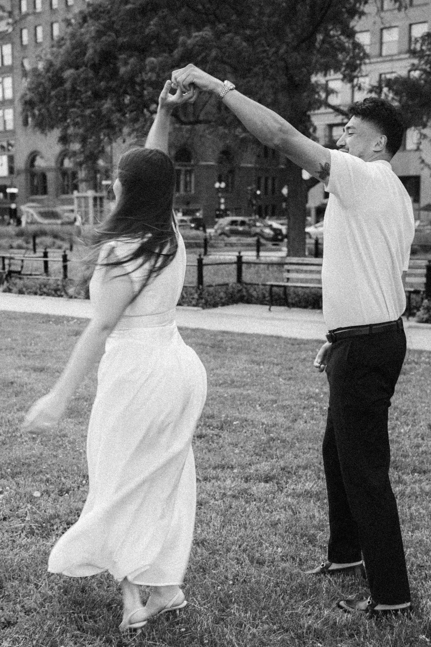 Black and white photo of couple dancing outdoors, man spinning woman in white dress on grass.
