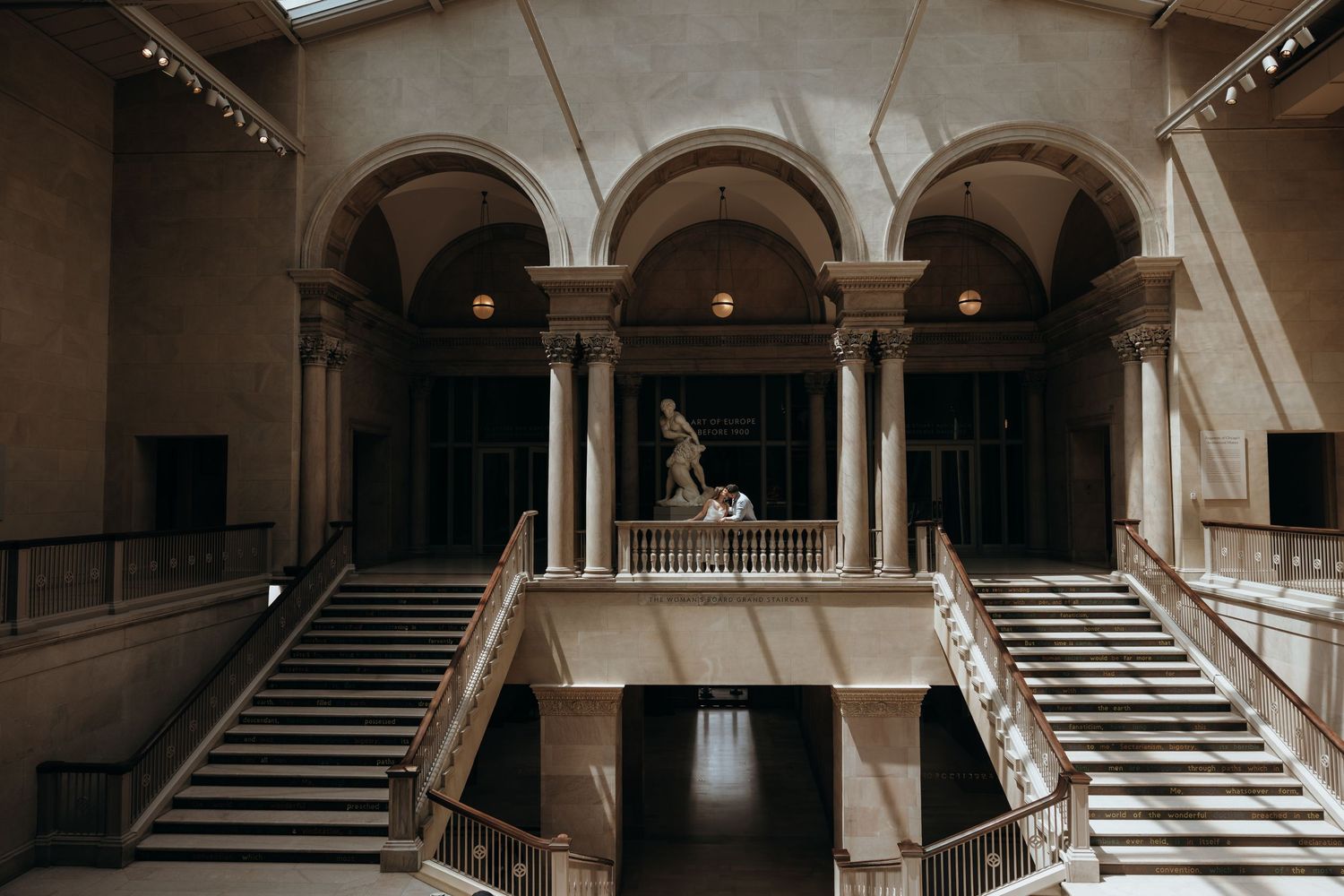 Elegant neoclassical atrium with dual staircases, arched colonnades, and a classical bronze statue centerpiece.