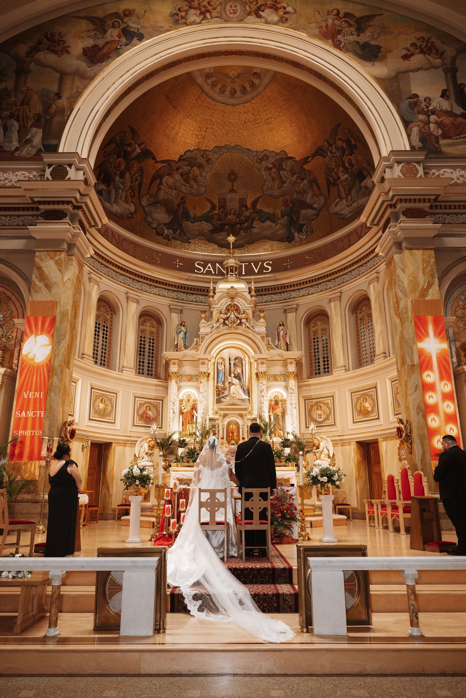 Bride and groom kneeling at ornate church altar during Catholic wedding ceremony beneath golden domed ceiling.