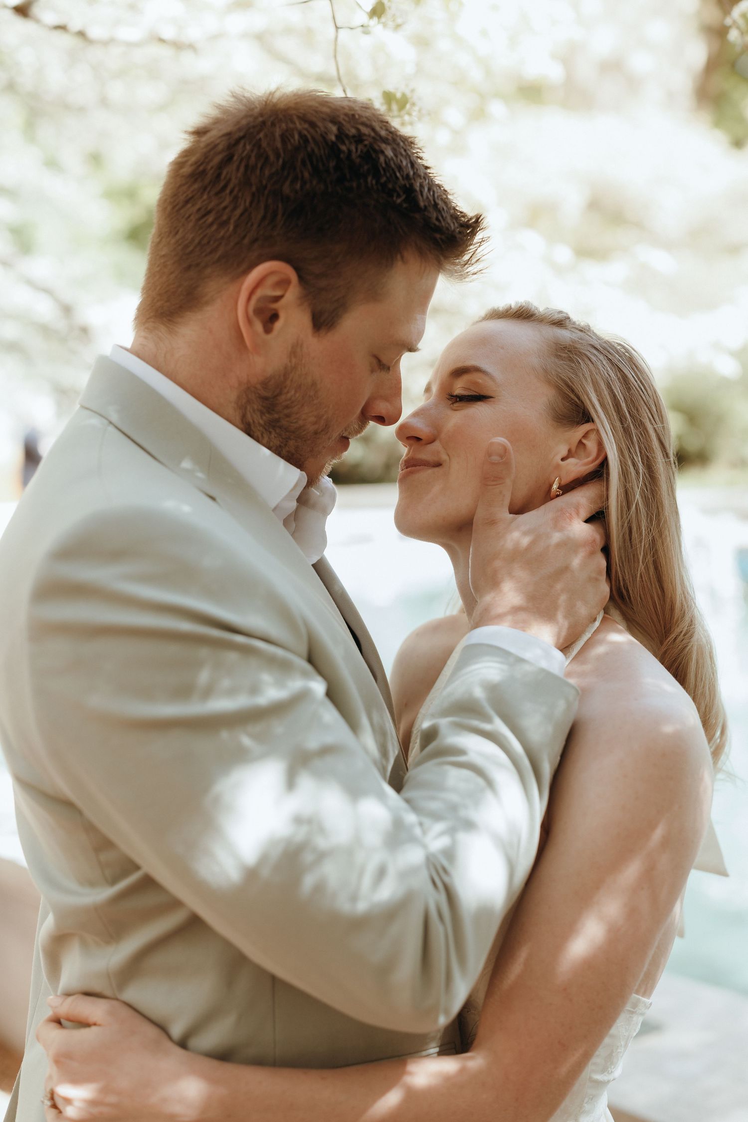 Groom gently cradles bride's face as they share a romantic moment under flowering trees.