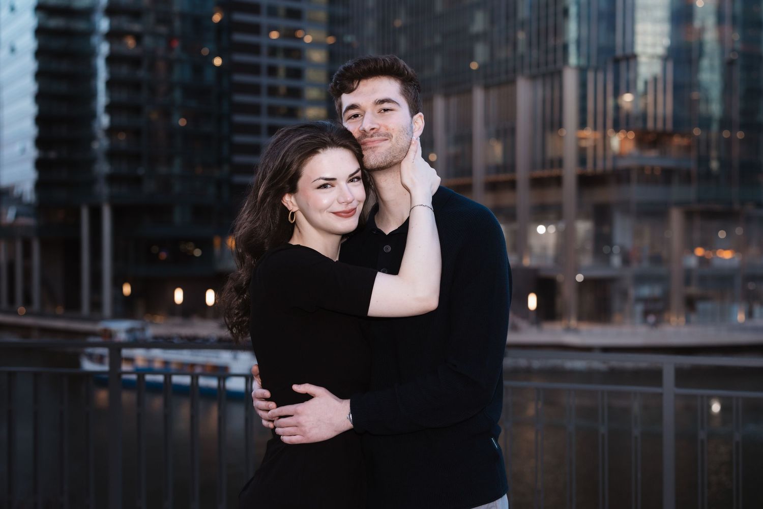 A couple poses together smiling warmly with the Chicago river and skyline glowing behind them.