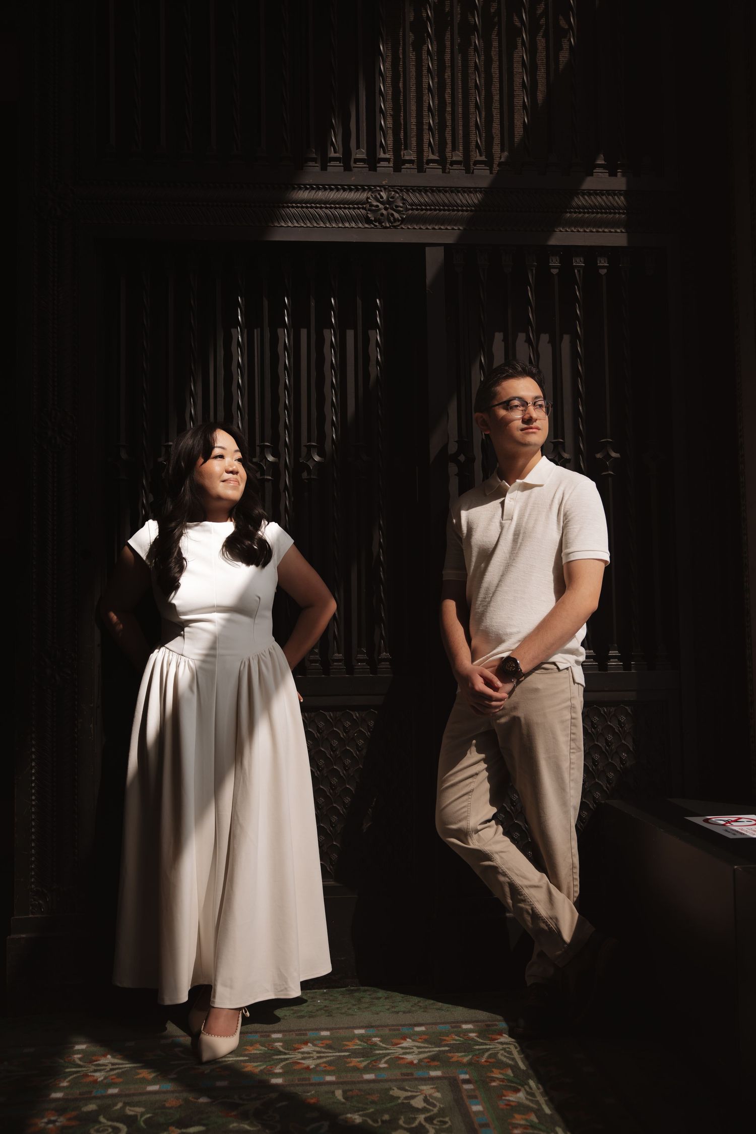 Elegant couple stands apart near dark ornate wooden architectural details.