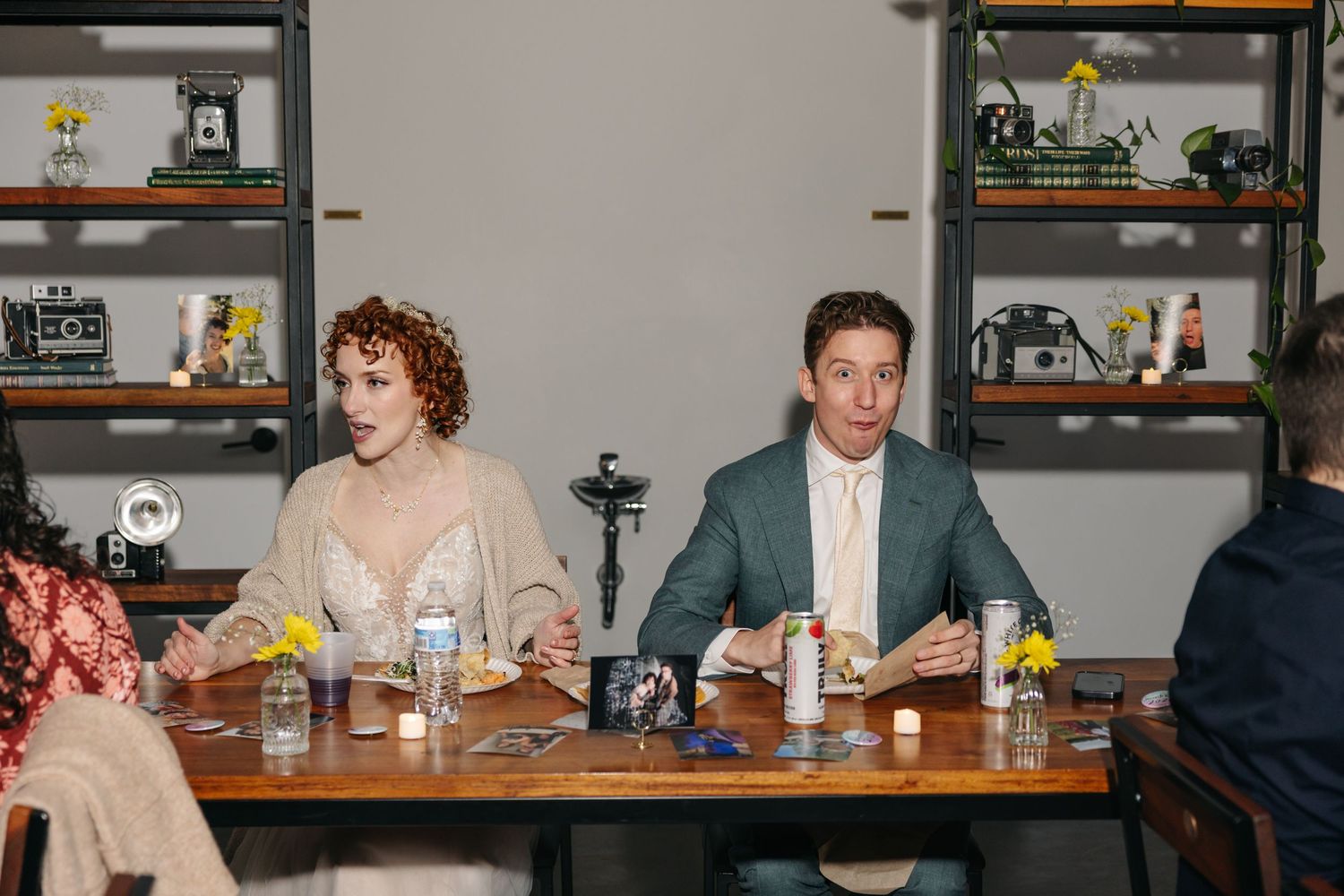 Bride and groom sit together at reception dinner table in modern industrial venue with shelving.