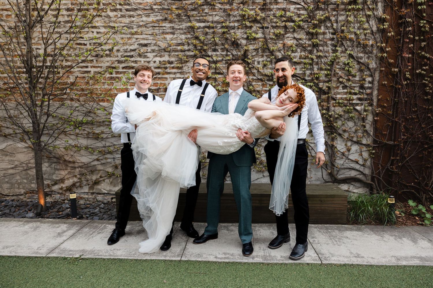 Groom and groomsmen in suspenders playfully carry bride in white wedding gown outside brick wall venue.
