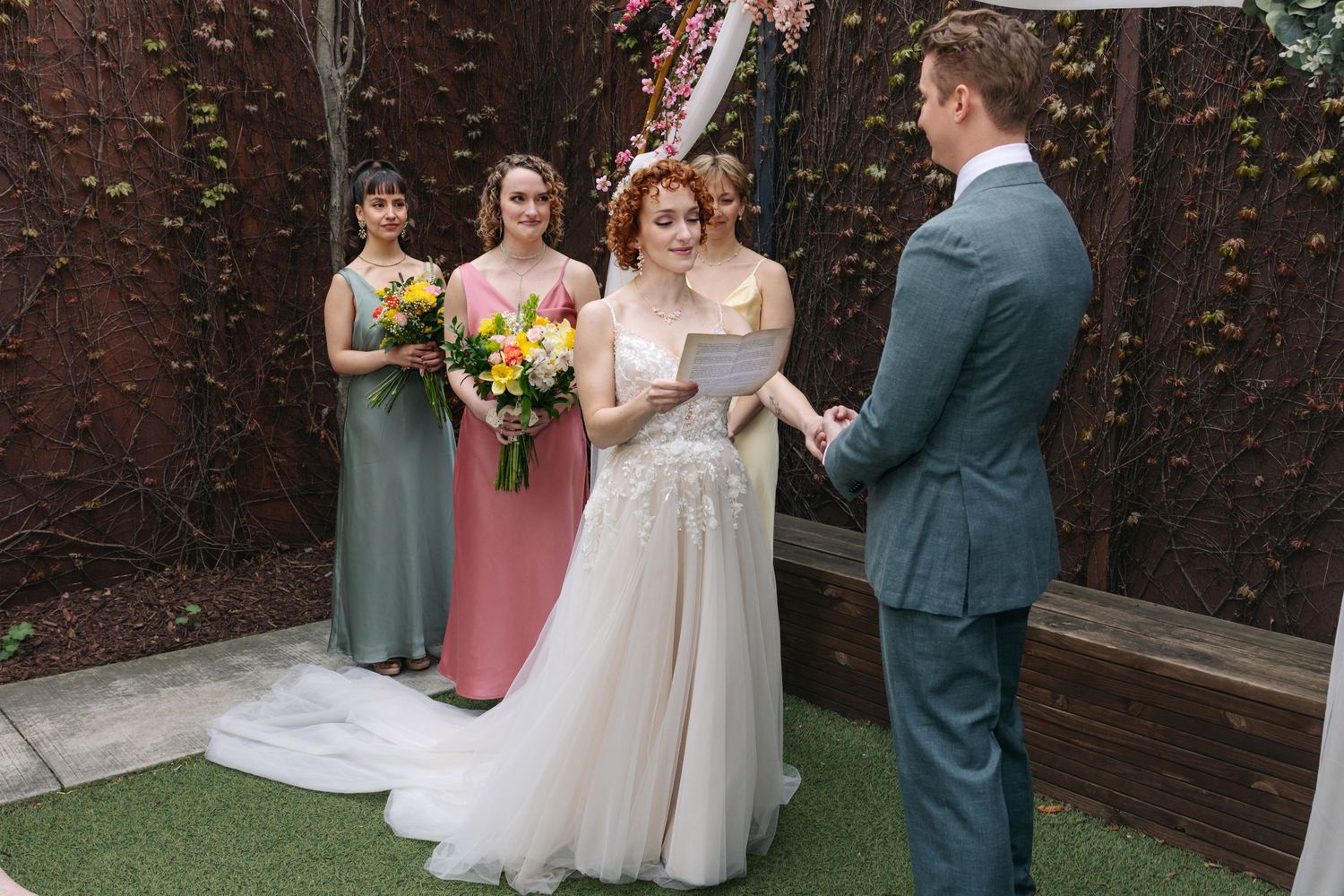 Bride reads vows to groom during outdoor wedding ceremony with bridesmaids in colorful dresses standing nearby.