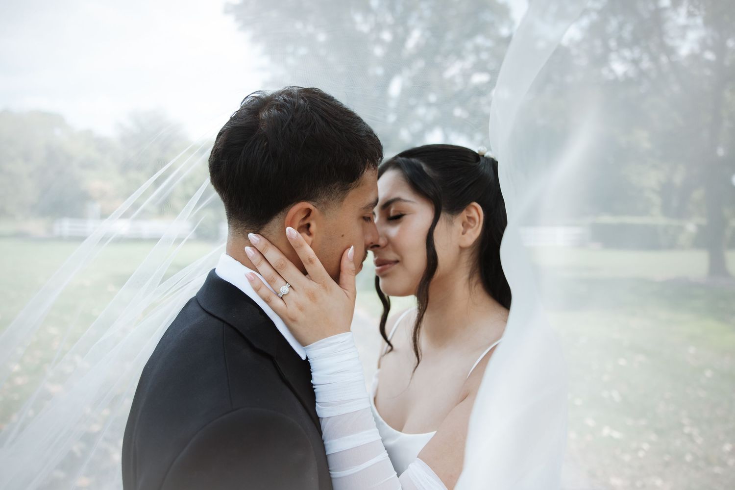 Bride and groom share an intimate moment under a flowing veil outdoors on their wedding day.