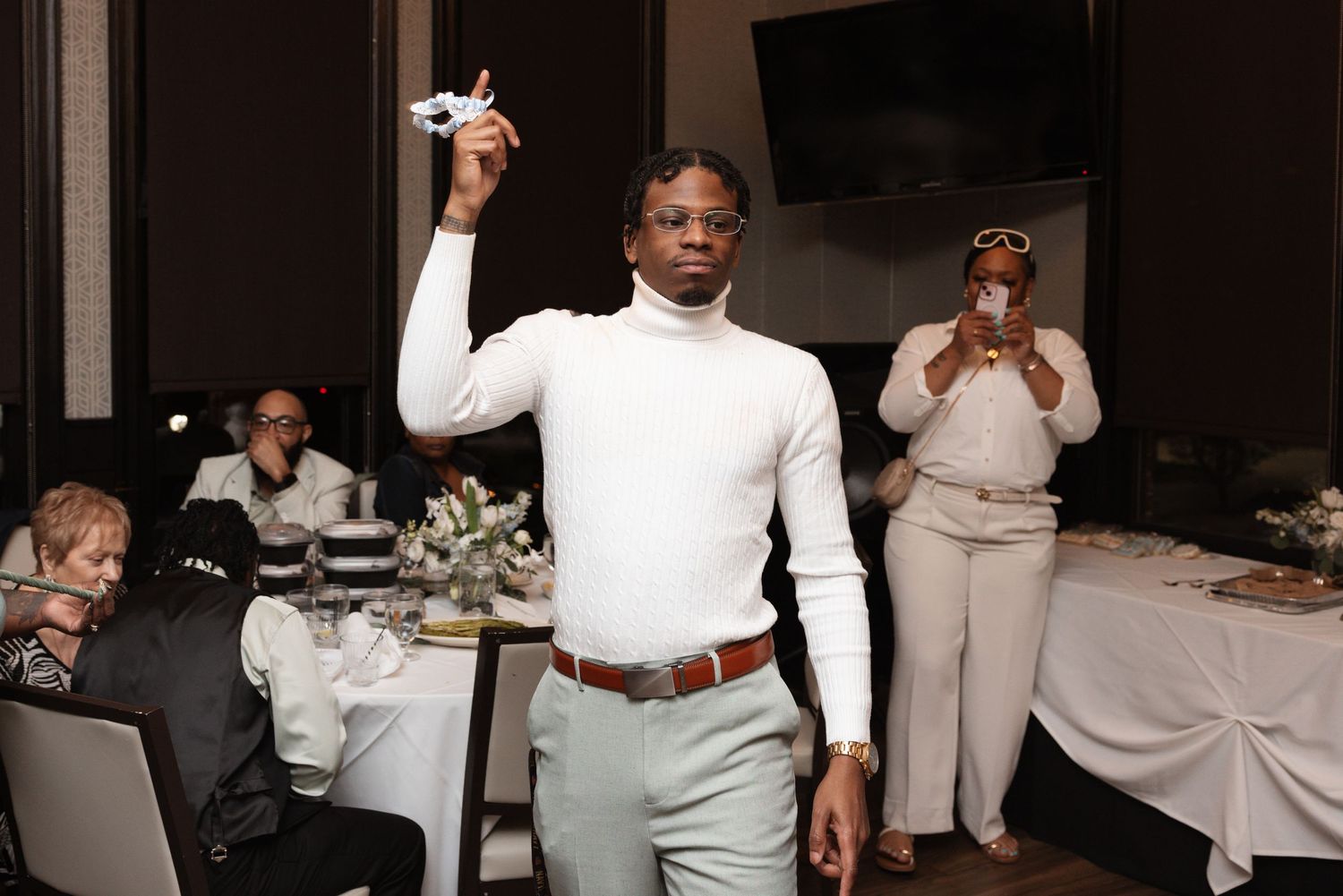 Man in white turtleneck holds toy airplane aloft while woman photographs him at wedding reception.