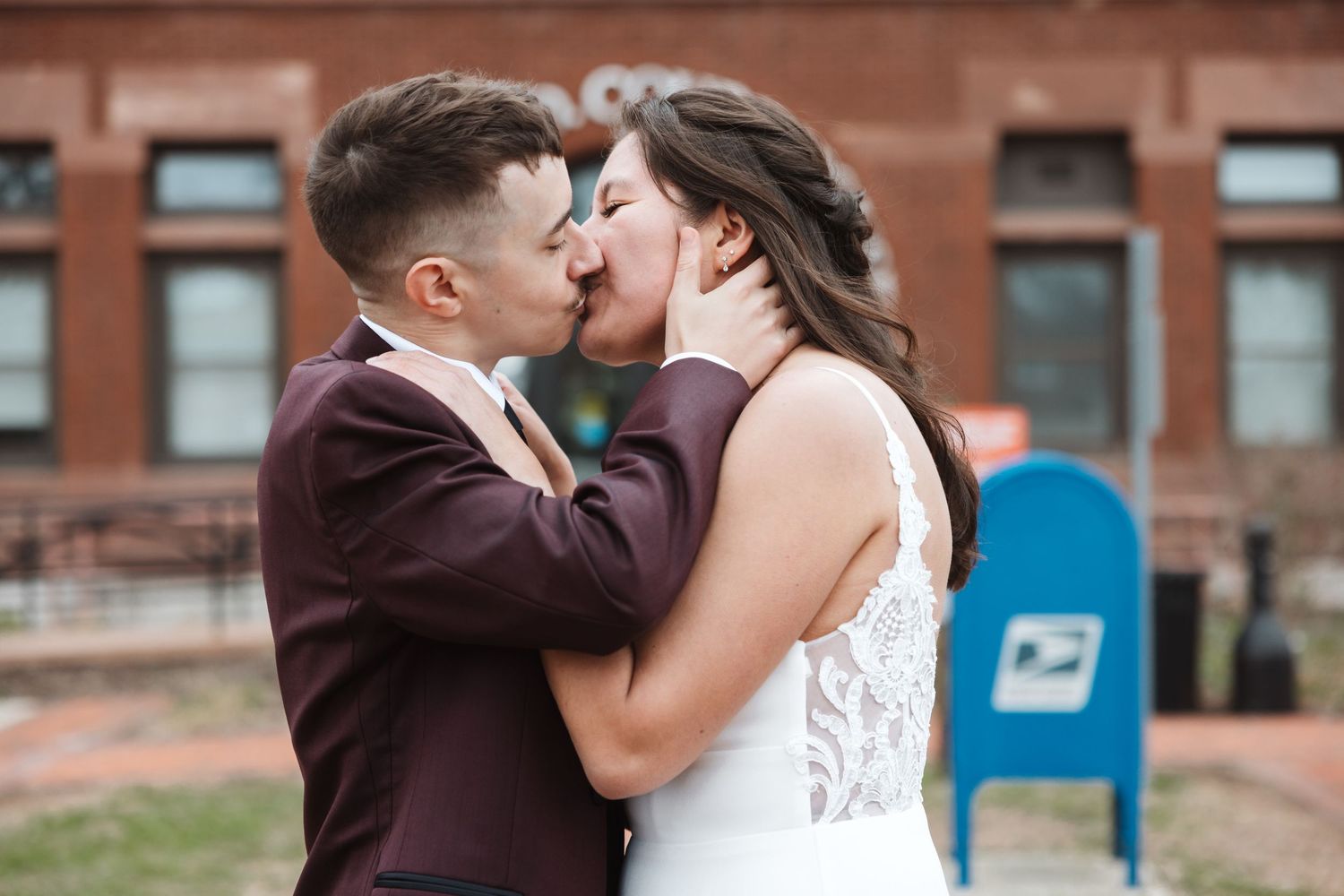 Couple sharing a romantic kiss in wedding attire outside a brick building, groom in burgundy suit, bride in white lace gown.
