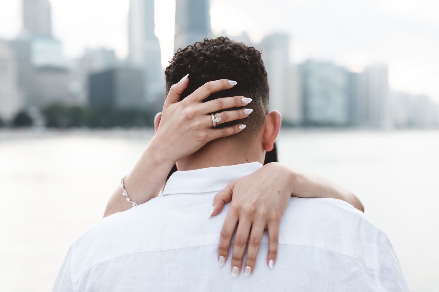 Close-up of woman's hands with engagement ring embracing man from behind near waterfront.