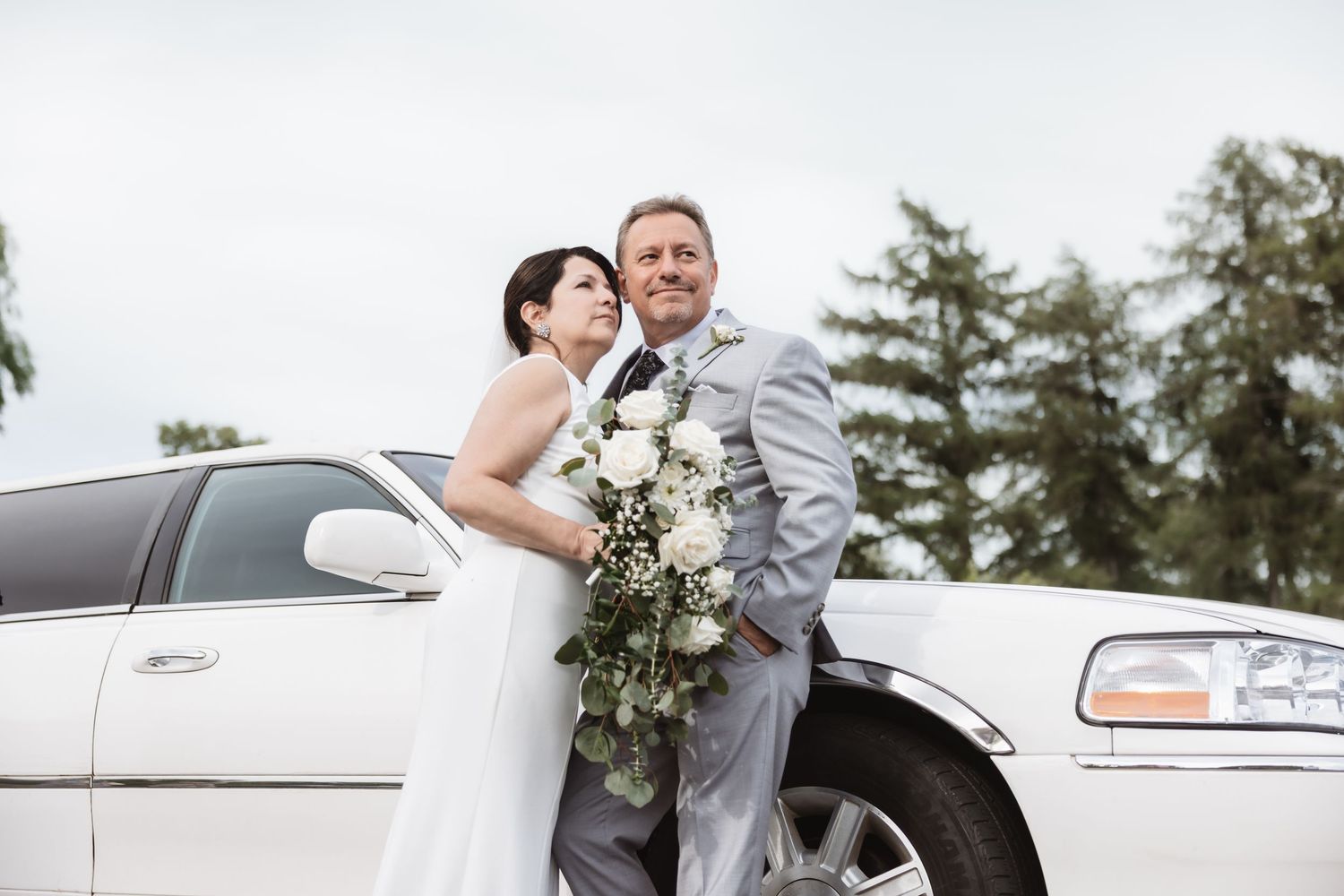 Bride and groom posing beside a white limousine, bride holding white floral bouquet, groom in gray suit.