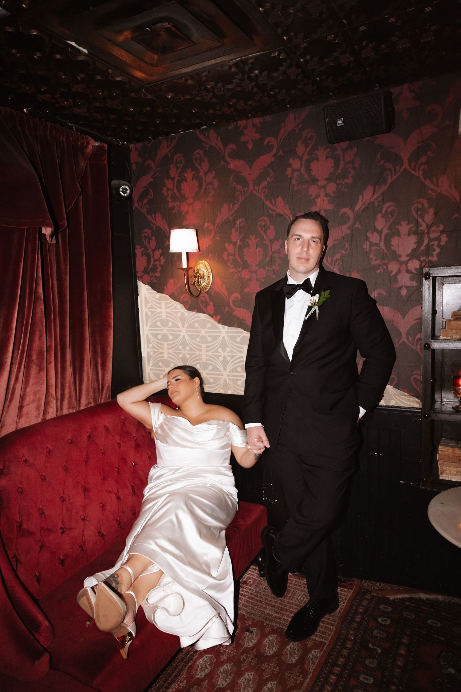 Bride lounges on red velvet sofa holding grooms hand in ornate decorated reception room.