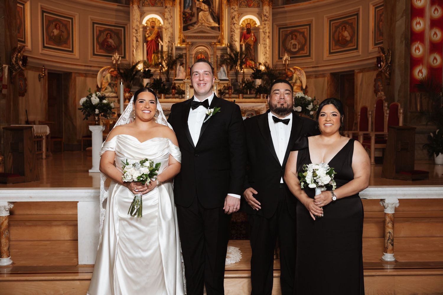 Wedding party of four posing at church altar, bride in white gown with groom and guests in formal black attire.