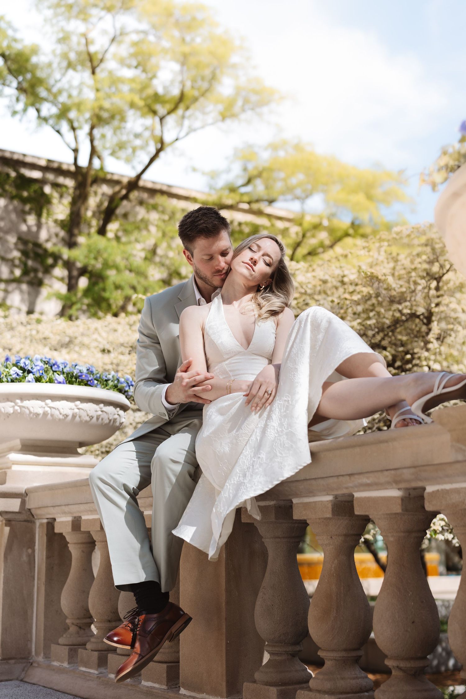 Bride in white lace dress reclines against groom on stone balustrade in a sunny garden setting.