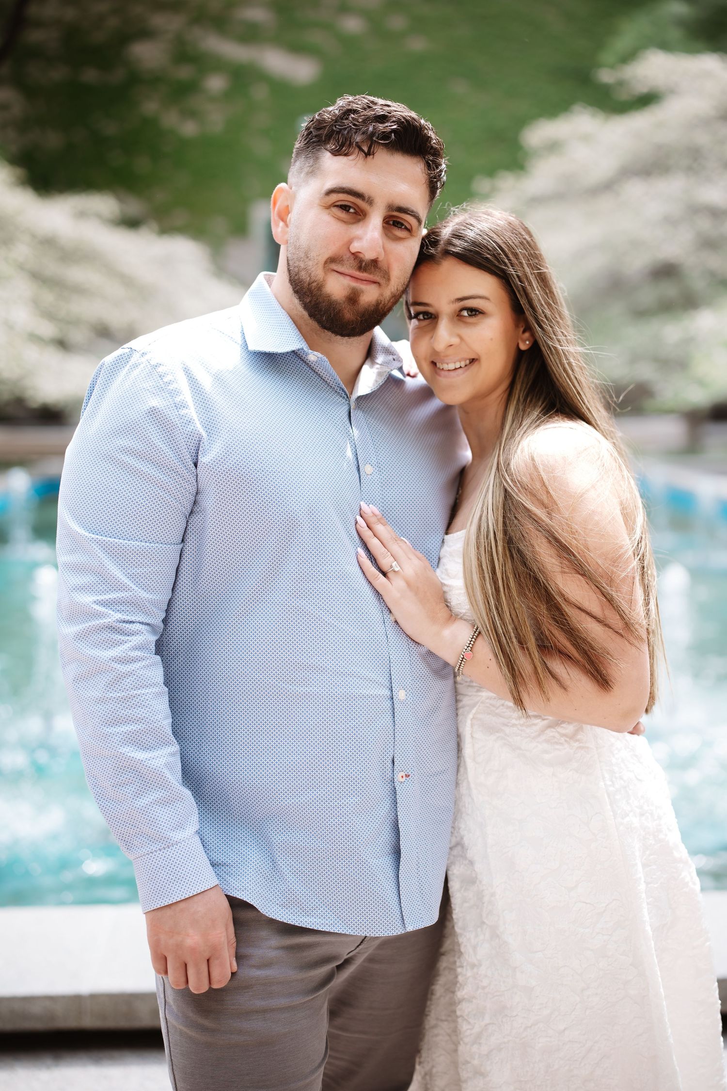 Couple posing outdoors near a fountain, woman in white dress leaning on man in blue shirt, surrounded by blooming trees.