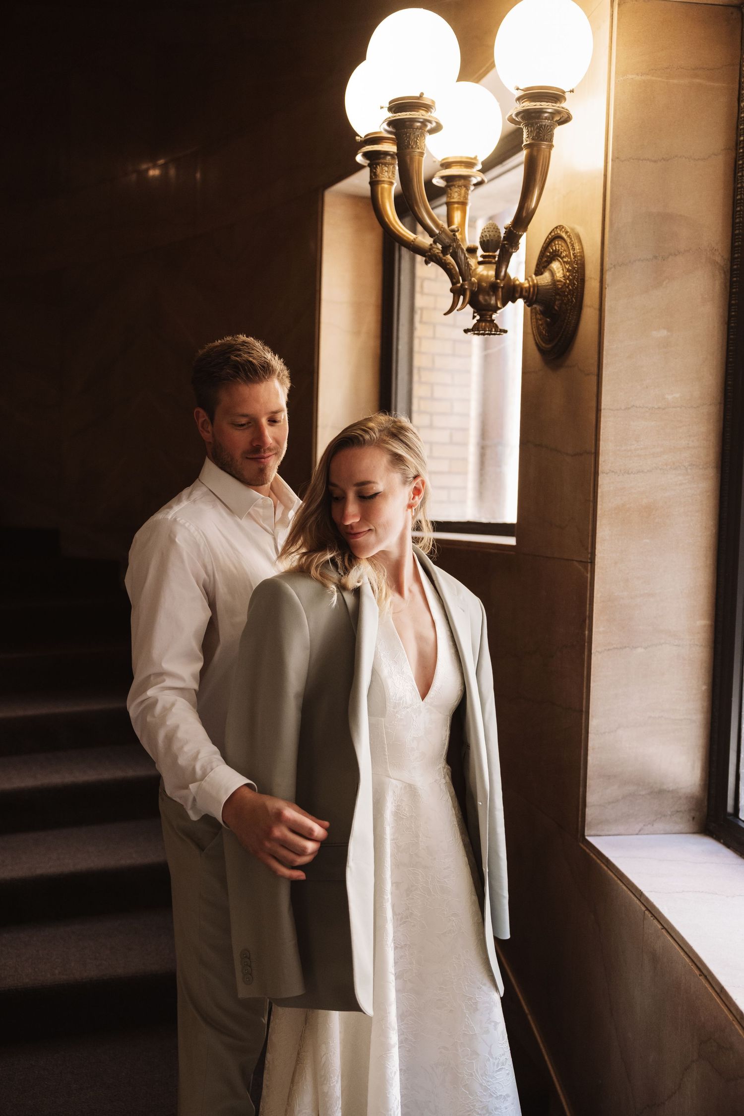 Groom helps bride into his suit jacket near a glowing wall sconce in an elegant interior.