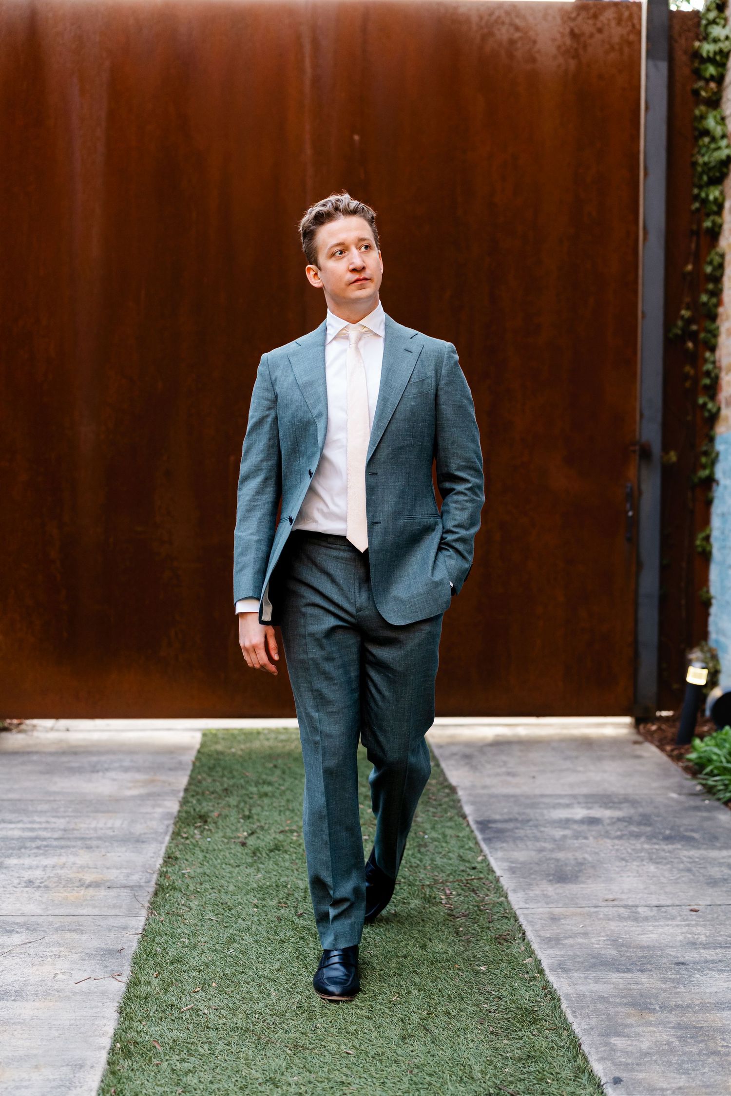 Groom in elegant teal suit walking confidently on grass path beside rustic metal wall outdoors.