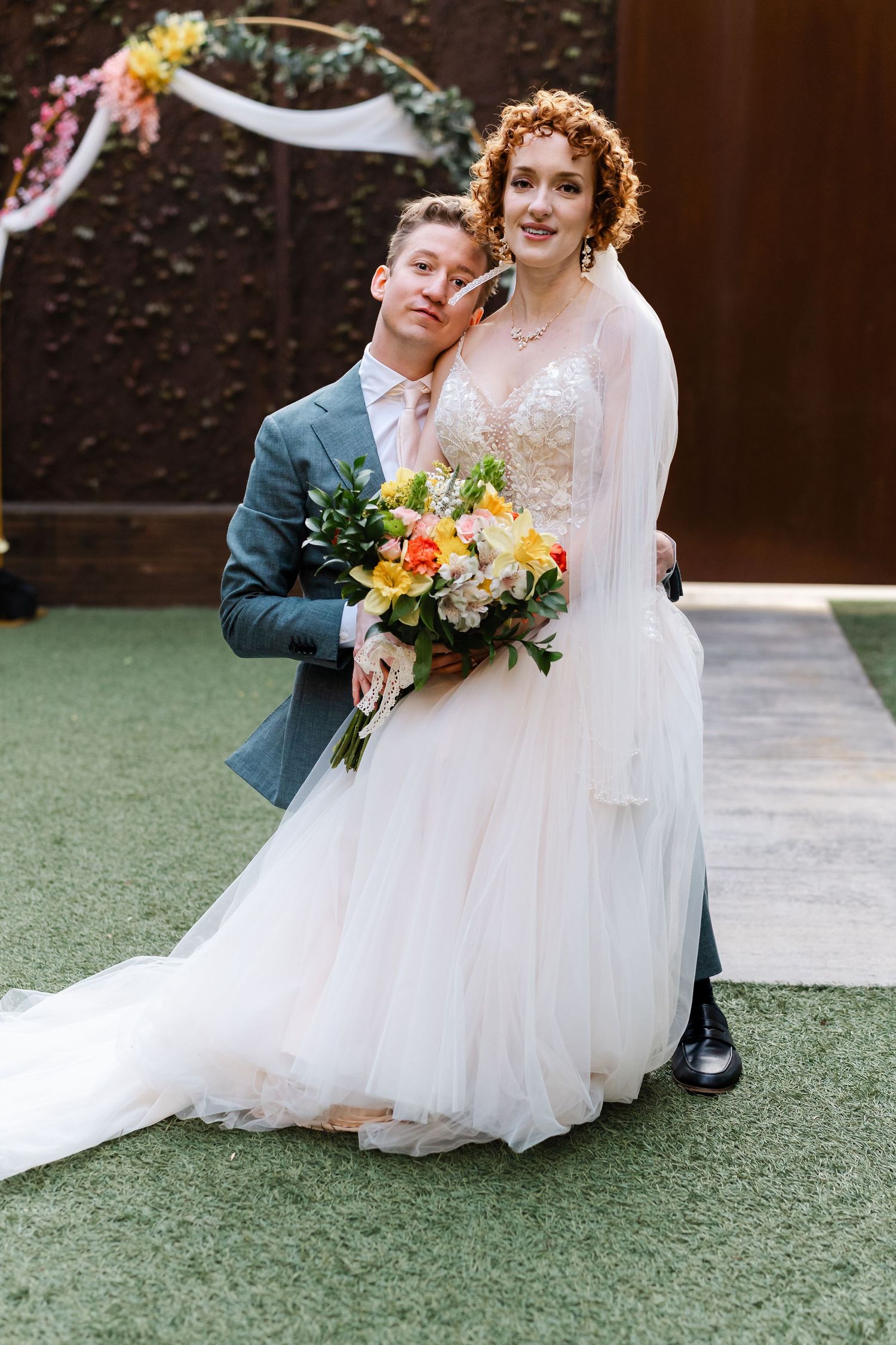 Bride in a layered tulle gown holds a colorful bouquet while groom in a gray suit leans close in front of a floral arch.