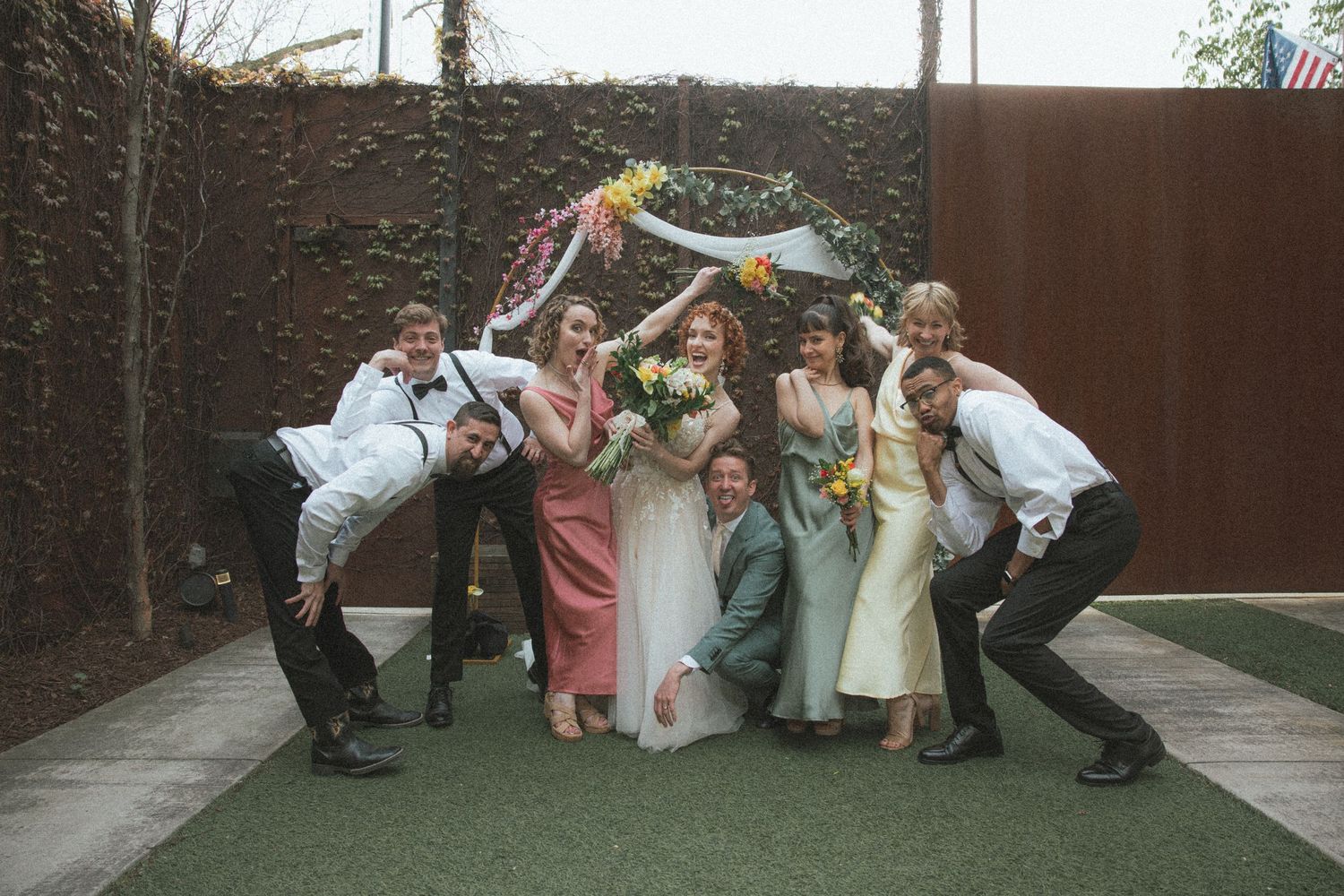 Wedding party poses playfully under floral arch at outdoor ceremony venue.