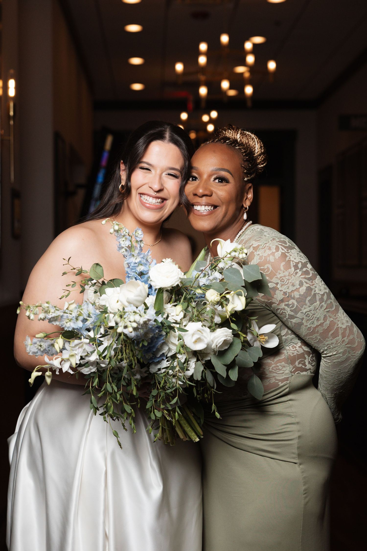 Smiling bride and bridesmaid in sage green lace gown pose together holding floral bouquets at reception.